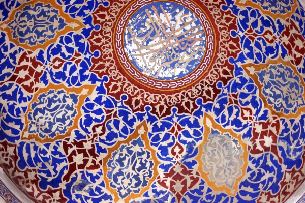 Red, blue, and gold intricate patterns on a white ground cover the inside of a dome in the Blue Mosque